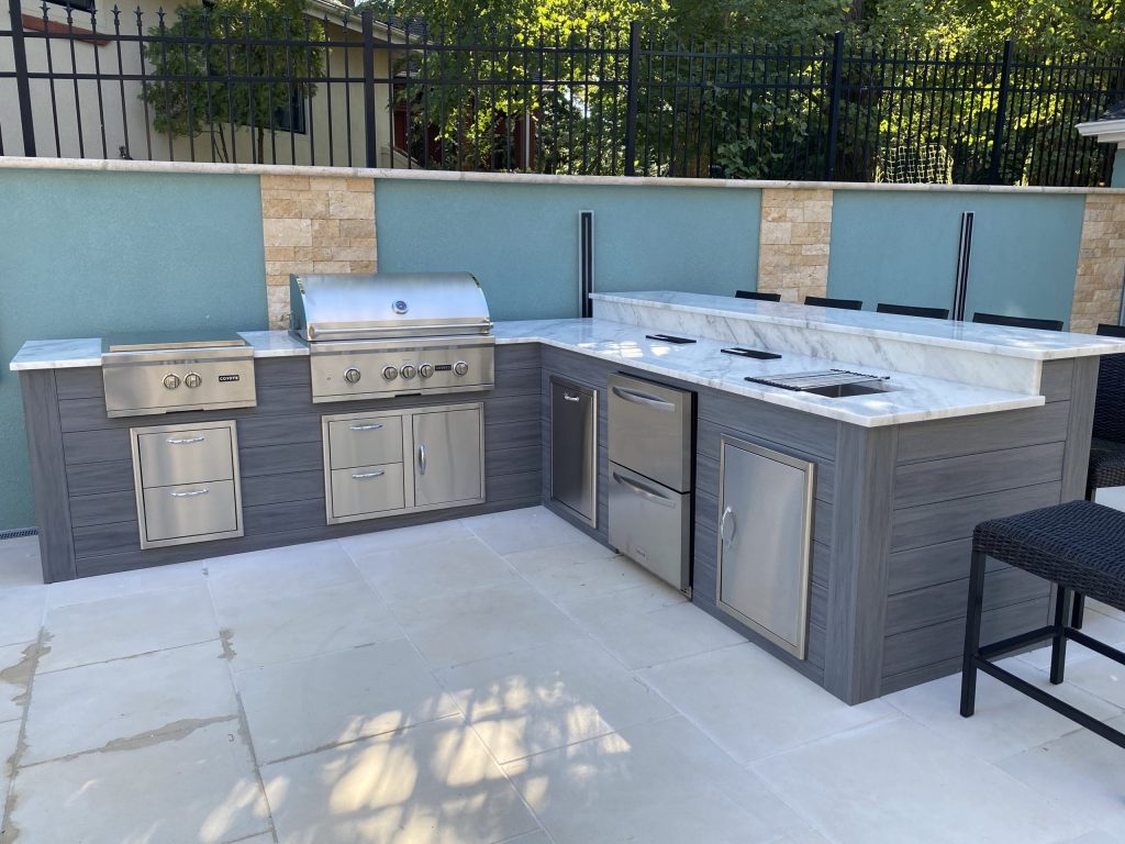 Outdoor kitchen with a stainless steel grill, side burners, and marble countertops along a teal wall.