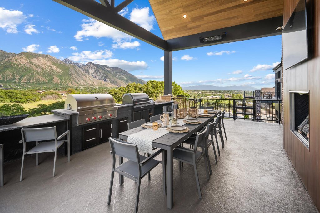 Outdoor covered patio with a long dining table set for eight, built-in grills, and mountain scenery in the distance under a blue sky.