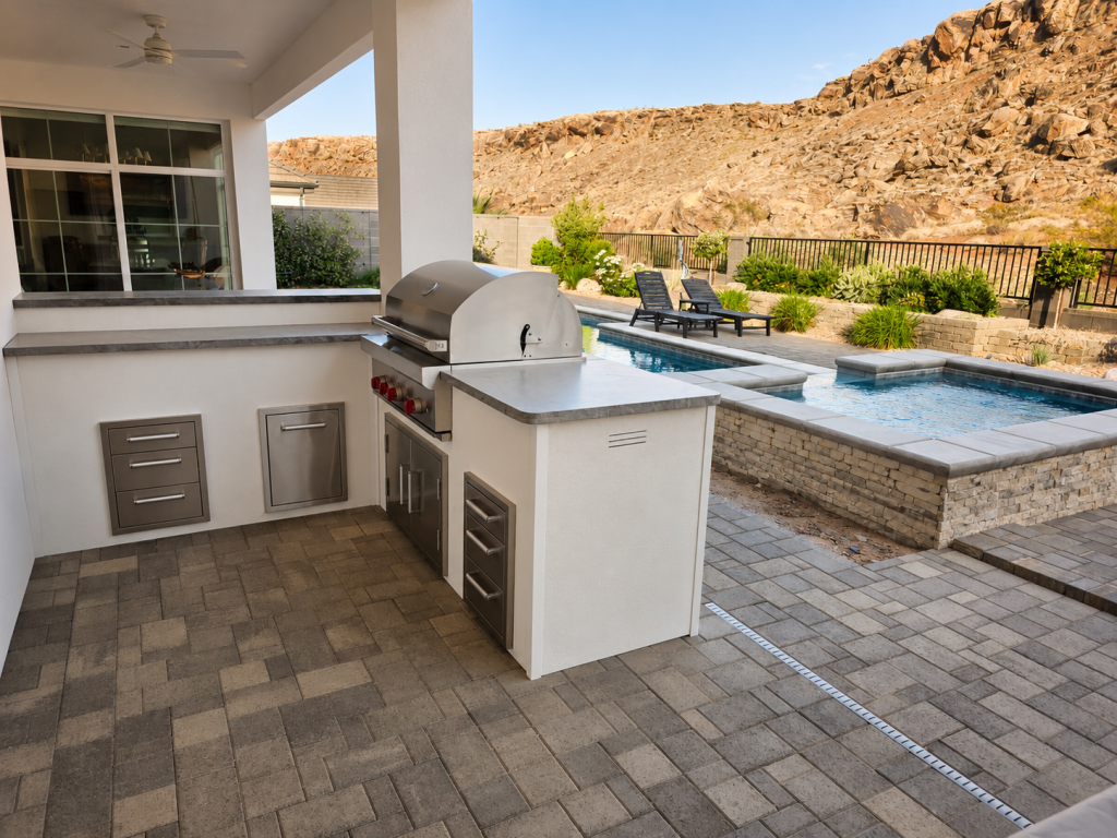 Outdoor stainless steel grill and counter area with built-in cabinets, next to a swimming pool and desert landscape in the background