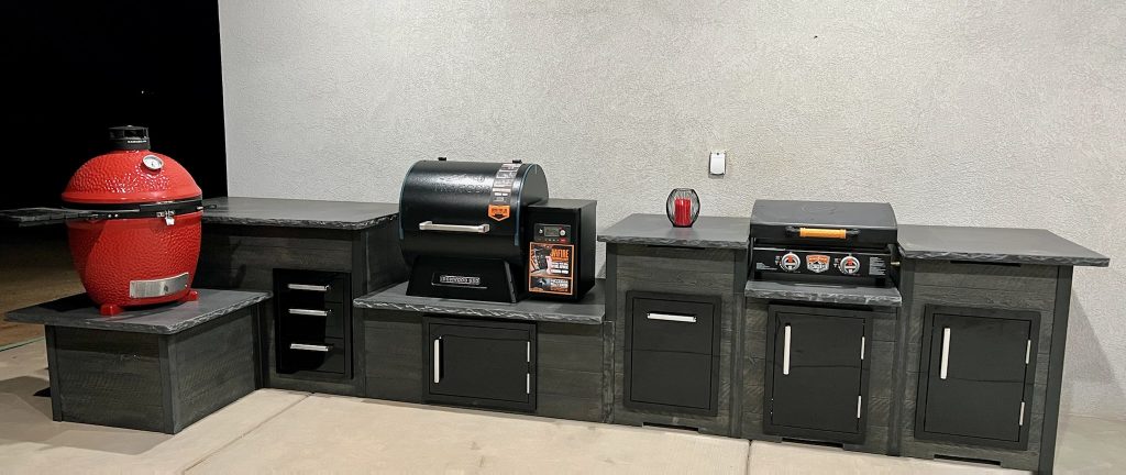 Outdoor kitchen setup with a red ceramic kamado grill on the left and a black pellet grill center, all on dark stone countertops with multiple cabinets and drawers nearby.