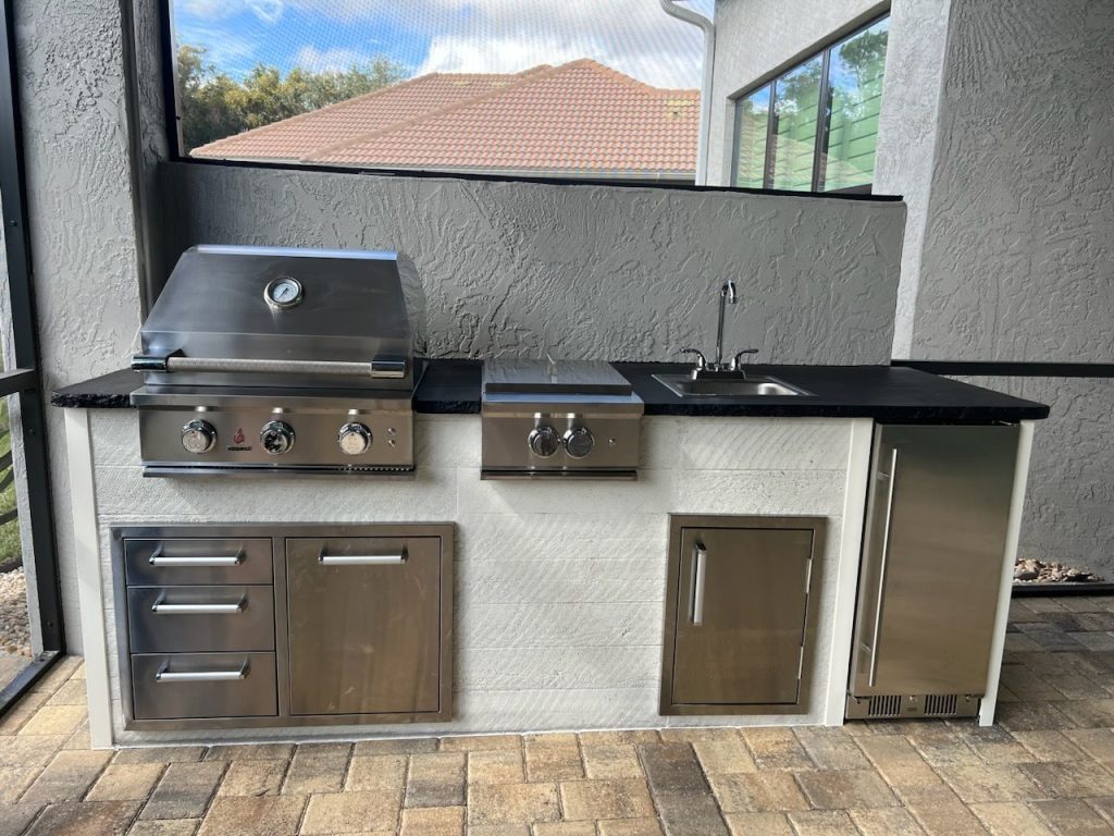 Outdoor stainless steel grill station with built-in thermometer, burners, and storage drawers under a dark countertop.