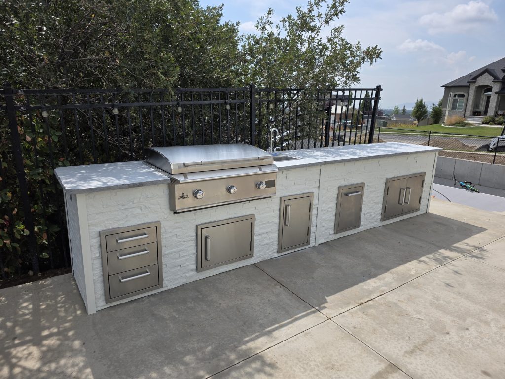 Outdoor stainless steel barbecue grill with storage drawers built into a white stone island, black iron fence behind, and a suburban house in the distance.
