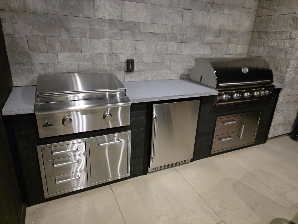 Outdoor stainless steel grill station with two built-in grills set against a textured stone wall and quartz countertop edge.