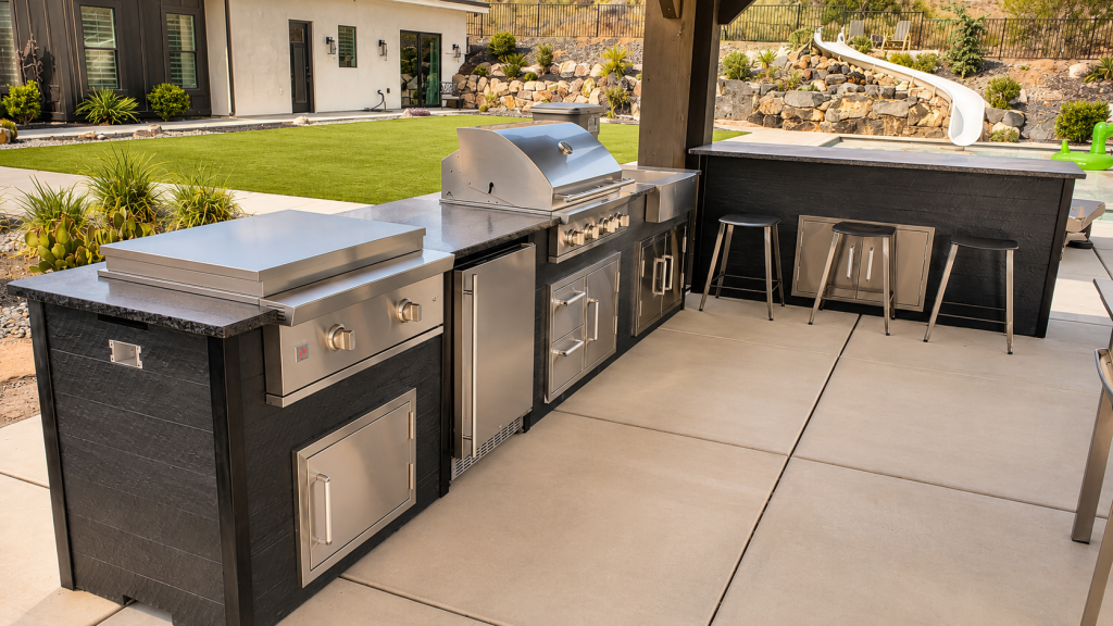 Modern outdoor kitchen with a stainless steel grill, side burners, fridge, and storage drawers under a covered patio, adjacent to a pool area.