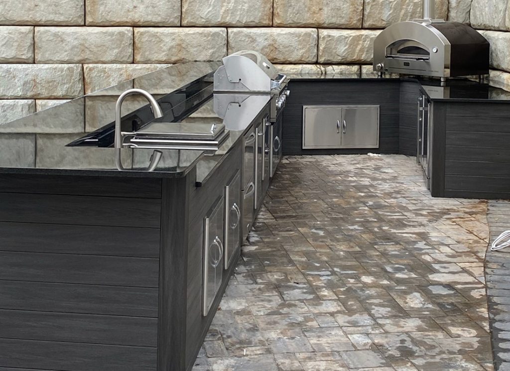 Outdoor stainless steel kitchen with grill, oven, and drawers along dark wood cabinets against a stone wall.