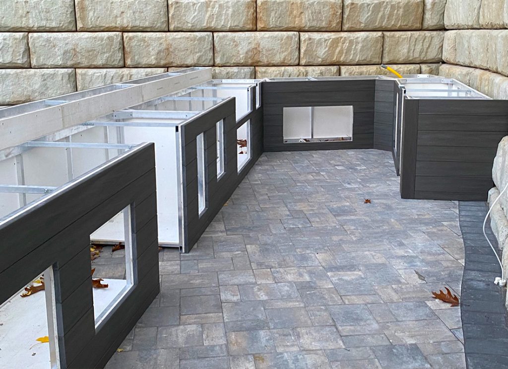 Partially assembled outdoor wall panels in dark gray with window cutouts on a gray paver patio, against a stone retaining wall.