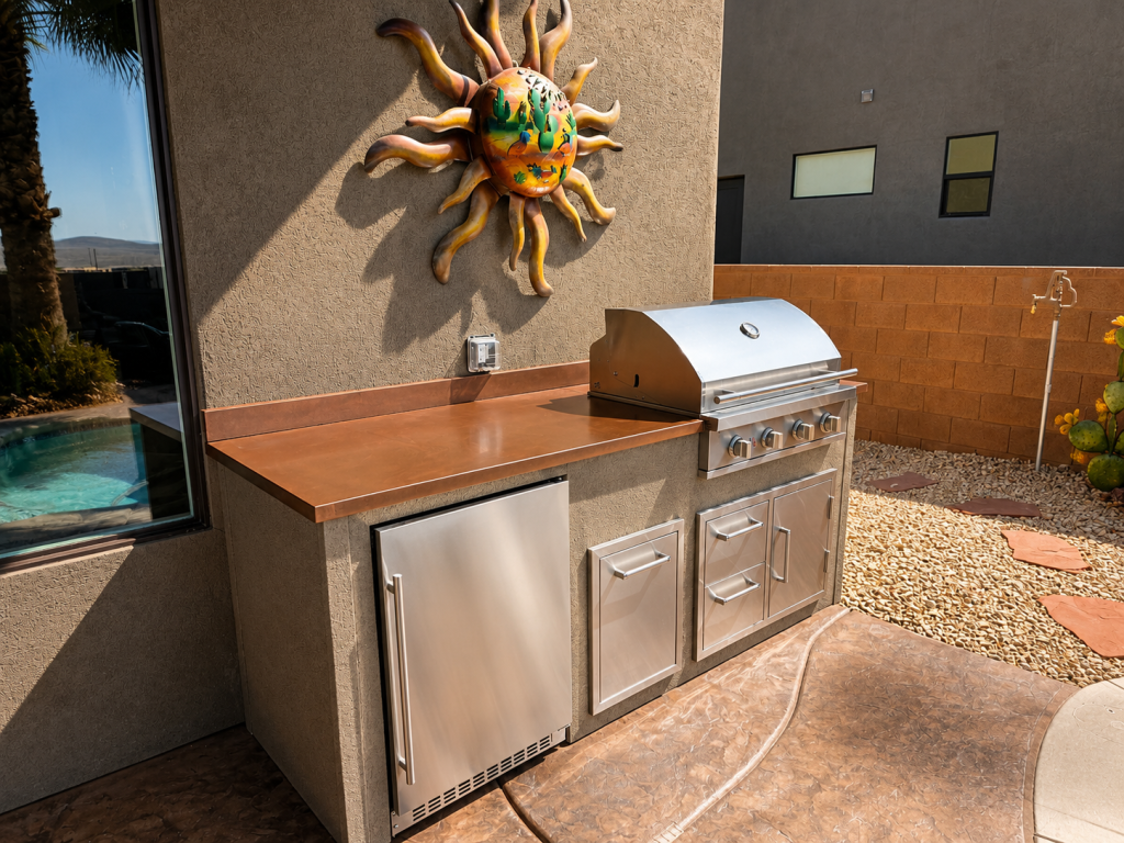 Outdoor stainless steel built-in grill with copper countertop and storage cabinets, sun sculpture on textured wall in an outdoor yard setting.