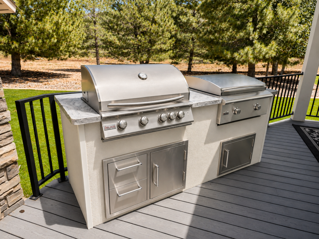 Two stainless steel gas grills with side counters and storage cabinets on a wooden deck, outdoors among trees.